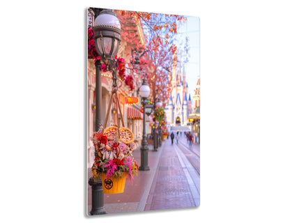 A festively decorated street in a theme park during fall, with colorful flower baskets hanging from lamp posts, autumn foliage on trees, and Cinderella's iconic castle in the background.
